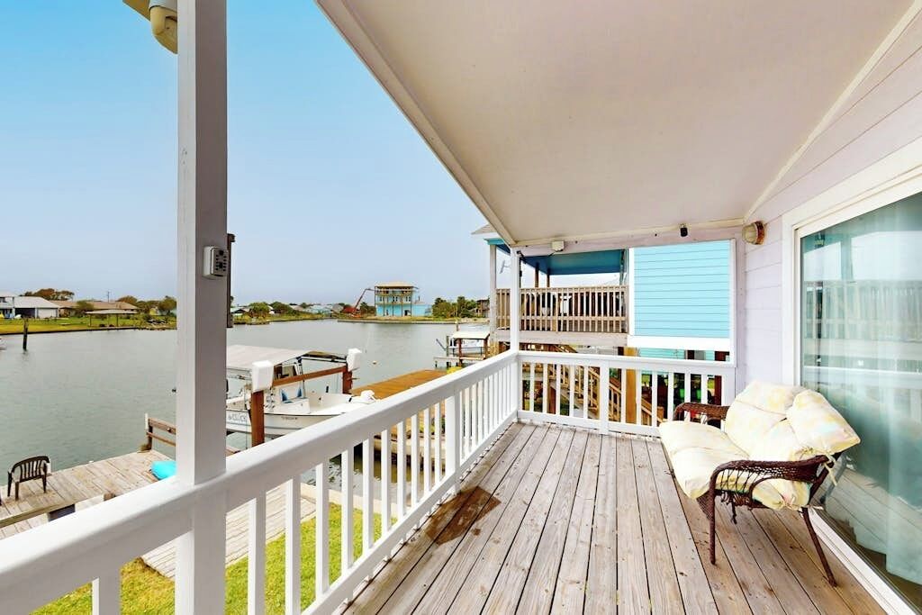 A wooden deck with a white railing, a covered seating area with cushions, and a view of a body of water and distant buildings.