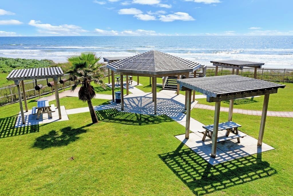A beachside gazebo with a view of the ocean, surrounded by picnic tables and a palm tree.