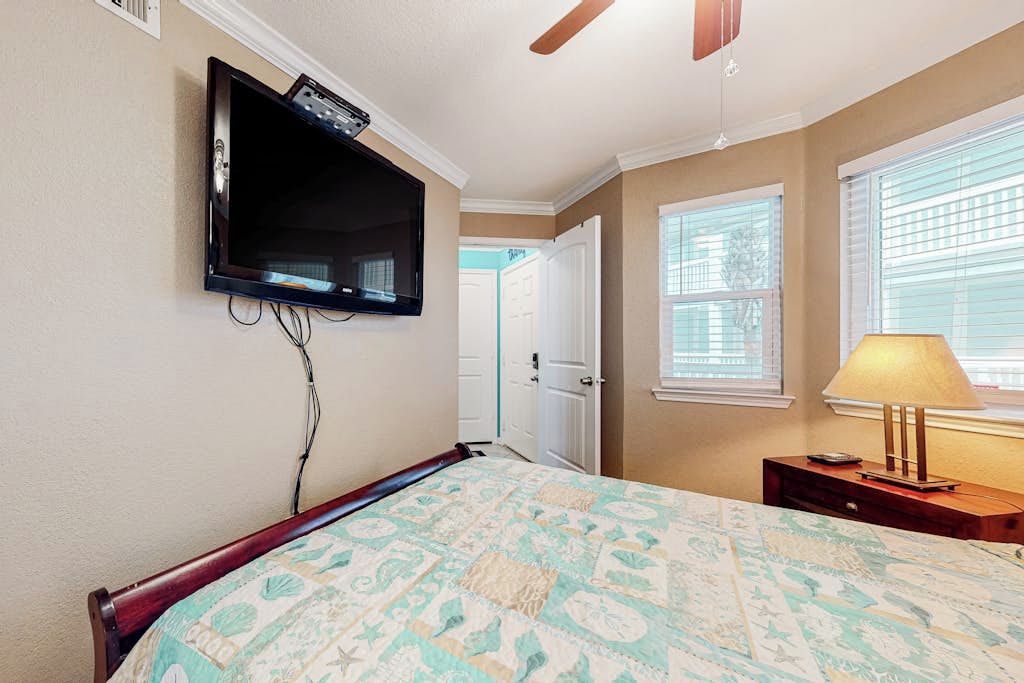 A bedroom with a bed, a television mounted on the wall, a ceiling fan, and a window with blinds.