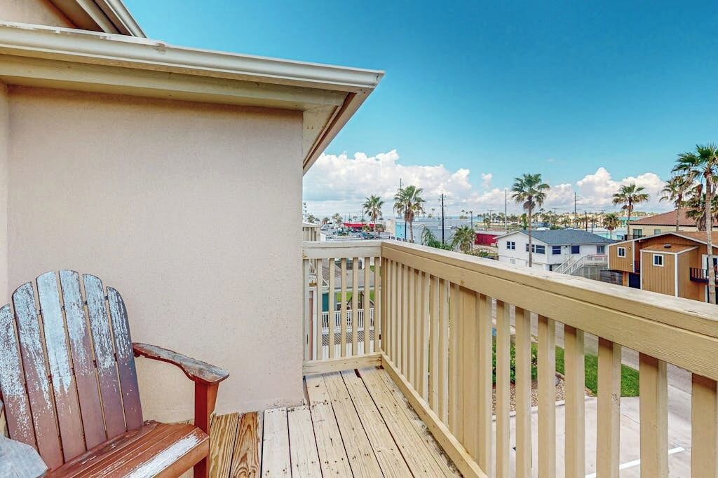 A wooden deck with a single Adirondack chair, a railing, and a view of a residential area with houses and palm trees.