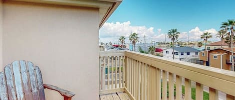 Una terraza de madera con una sola silla Adirondack, barandilla y vista a una zona residencial con casas y palmeras.