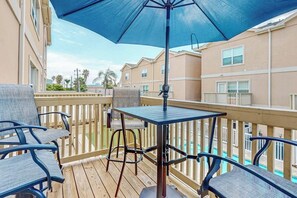 A balcony with a table and chairs, a blue umbrella, and a view of a swimming pool. - Star Beachside Condominiums 106 (South Padre Island)