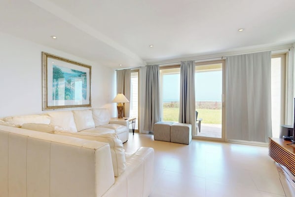 Un salón espacioso con un sofá blanco, una mesita de noche con una lámpara y un televisor. Amplias ventanas con cortinas, y vistas al exterior.