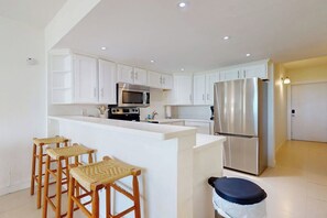 A modern kitchen with white cabinetry, a stainless steel refrigerator, a microwave, and a bar area with stools. - Solare Garden Villa 220 (South Padre Island)