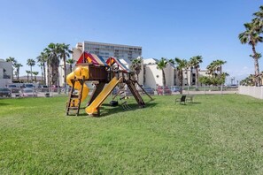 A playground with a slide and climbing structures, surrounded by a fenced area with palm trees and a building in the background. - Solare Garden Villa 220 (South Padre Island)