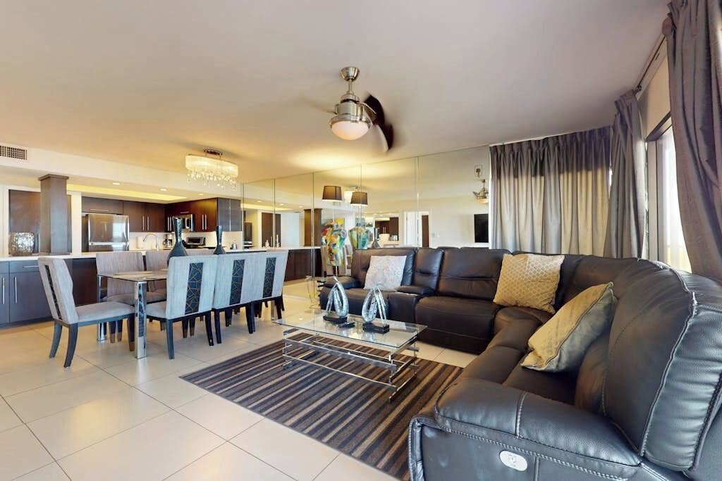 A modern living room with a black leather sofa, a dining area with a glass table, and a kitchen in the background.