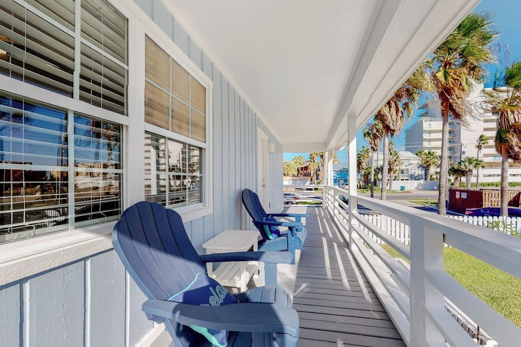A covered outdoor seating area with chairs and tables, overlooking a street with palm trees and buildings.