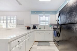 A kitchen with white cabinets, a black refrigerator, a window with blinds, and a whiteboard on the wall. - Lazy Seagull Beach House - Upstairs (South Padre Island)