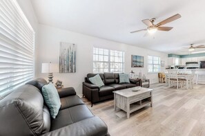 A spacious living area with a sofa, coffee table, and a dining table with chairs. There are ceiling fans, a kitchen area with cabinets, and a large window providing natural light. - Lazy Seagull Beach House - Upstairs (South Padre Island)