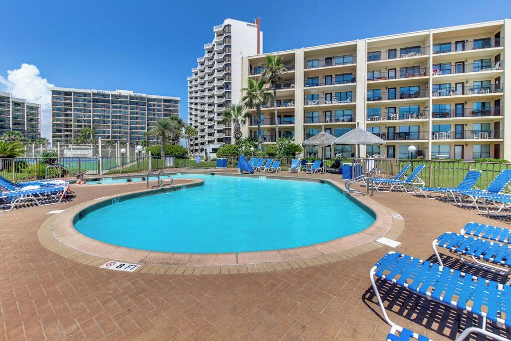 A hotel pool area with a clear blue pool, sun loungers, and a multi-story building in the background.