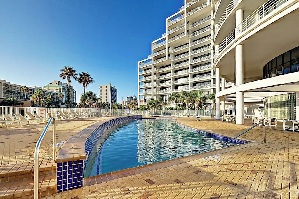 Ein Hotelpoolbereich mit einem geschwungenen Pool, Palmen und einem mehrstöckigen Gebäude mit Balkonen.