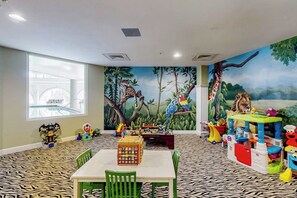A playroom with a jungle-themed mural, a table with chairs, and various children's toys. - Diamond Beach Resort - A Shore Thing (Galveston)