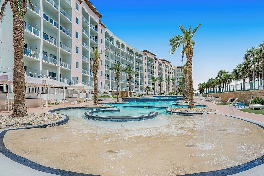 A hotel with multiple floors, a swimming pool with a fountain, and palm trees.
