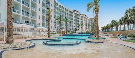 A hotel with multiple floors, a swimming pool with a fountain, and palm trees.