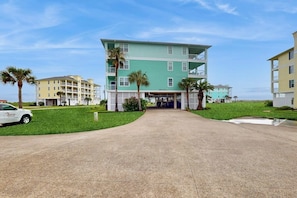A multi-story building with a flat roof, a parking area with a white car, and palm trees. - Pointe West Resort - Sand Swept (Galveston)