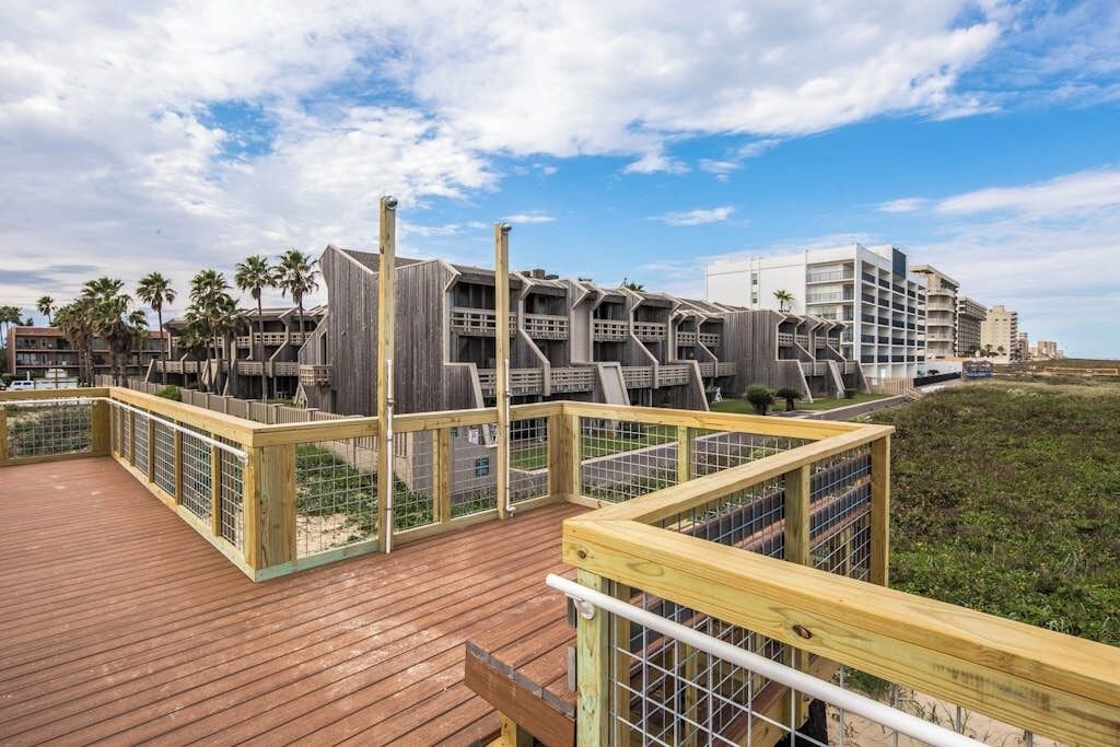 A wooden deck with a view of a modern building with multiple balconies, surrounded by palm trees and a clear sky.