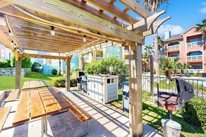 An outdoor patio with wooden benches and a grill area, surrounded by greenery and buildings. - Dawn Condominiums - Break of Dawn (Galveston)