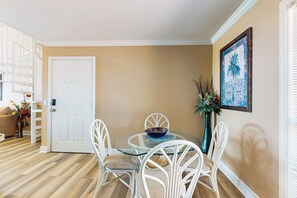 A dining area with a glass table and white chairs, a white door, and a wall-mounted painting. - Dawn Condominiums - Break of Dawn (Galveston)