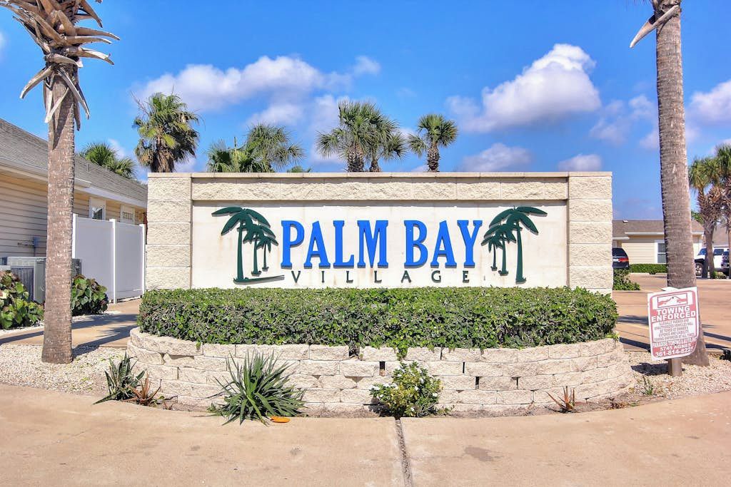 Une enseigne indiquant l’entrée de Palm Bay Village, avec des palmiers et un ciel dégagé.