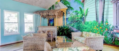 Une pièce meublée de meubles en osier, dotée d’un ventilateur de plafond, ornée d’une fresque représentant un paysage tropical et comprenant un vélo.