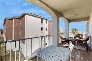 A balcony with a white table and wicker chairs, overlooking a building with brick and white walls. - Padre Island 104 - Ocean Views, Walk to the Beach (Corpus Christi)