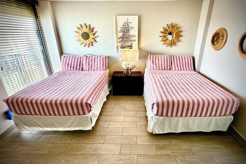 A bedroom with two beds, a window with blinds, and wall decor including sunburst mirrors and a framed picture.