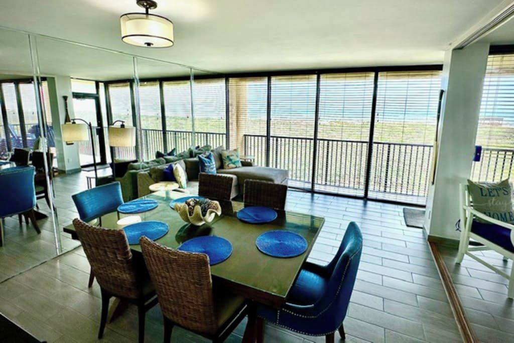 A dining area with a large table, chairs, and a view of the outdoors through large windows.