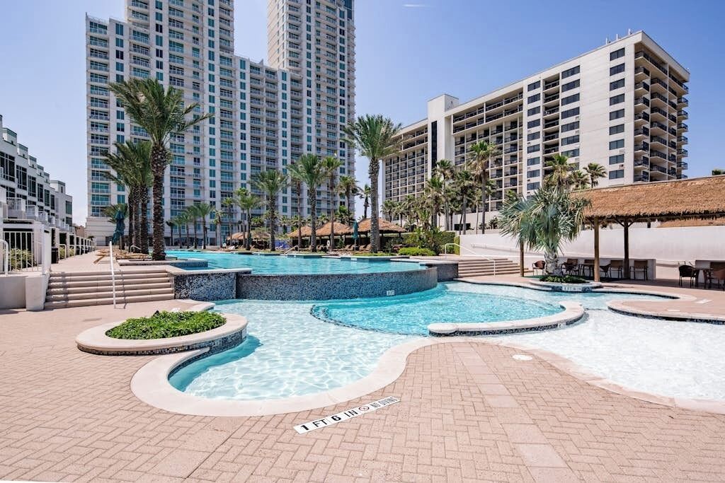 A hotel pool area with multiple swimming pools, palm trees, and high-rise buildings in the background.