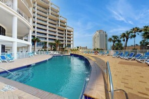A swimming pool with lounge chairs, a multi-story building with balconies, and palm trees. - Sunchase IV #517 - Wraparound Balcony & Pools (South Padre Island)