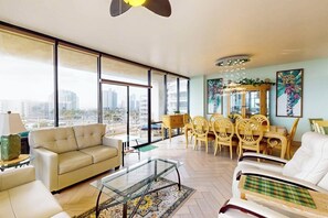 A spacious living room with a large window, a sofa, a coffee table, and a dining area with chairs and a table. - Sunchase IV #517 - Wraparound Balcony & Pools (South Padre Island)