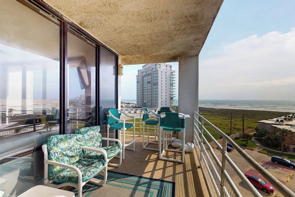 A balcony with a view of the ocean, a high-rise building, and a parking area.