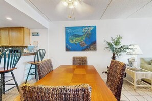 A dining area with a wooden table and woven chairs, a kitchen with wooden cabinets, and a wall-mounted painting of a mermaid. - Suntide II Condominiums #108 (South Padre Island)