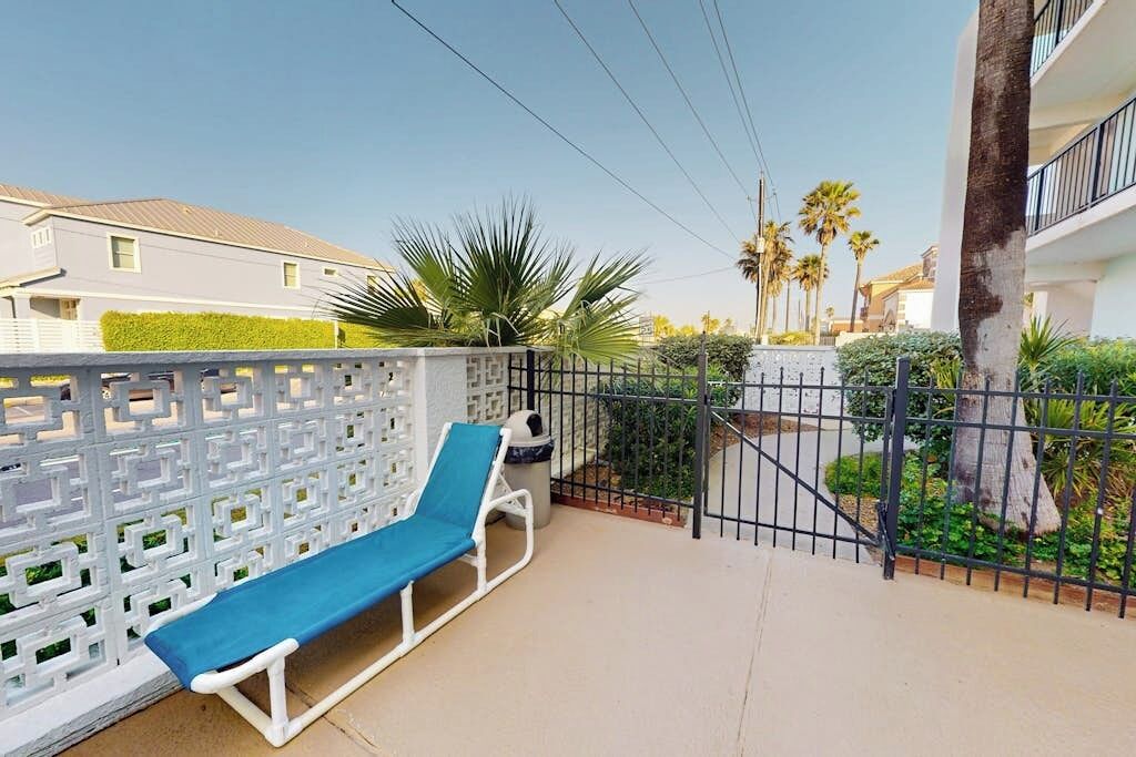 Un balcon avec une chaise blanche et un coussin bleu, une clôture blanche et une vue sur des palmiers et un immeuble.