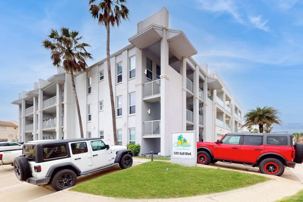 A multi-story residential building with balconies, a sign indicating it's a condominium, and two vehicles parked in front.