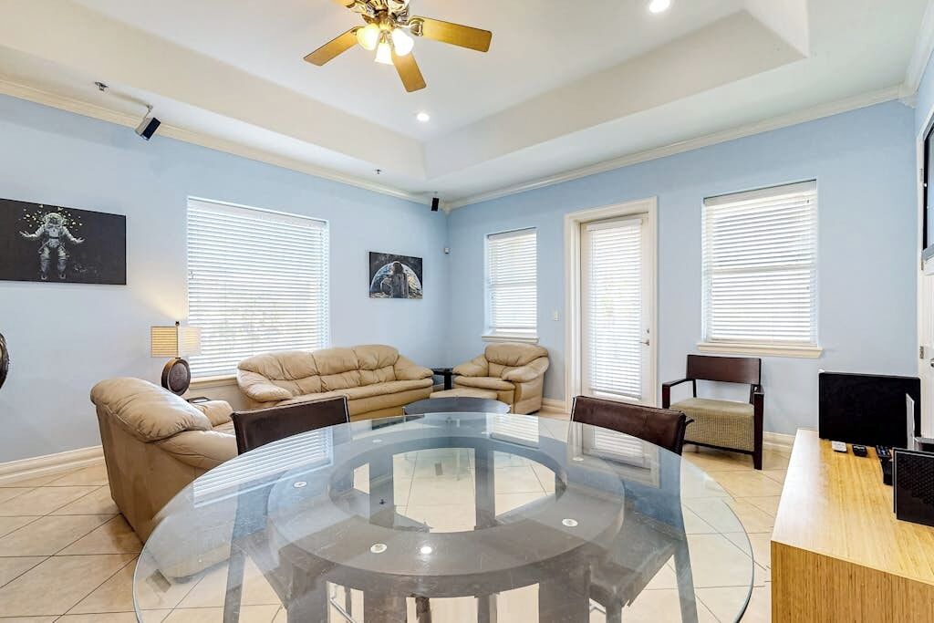 A room with a glass-top table surrounded by chairs, a ceiling fan, and a desk with a computer.