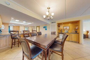 A spacious dining area with a large wooden table and multiple chairs, a kitchen area with wooden cabinets, and a chandelier. - Saida Towers IV #4906 (South Padre Island)