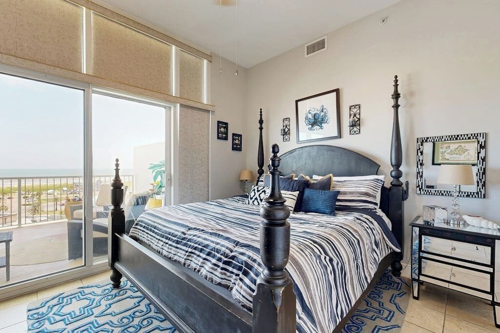Un dormitorio con un ventanal grande, una cama con cabecera y una mesita de noche.