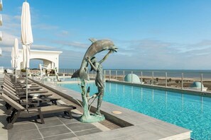 Una piscina con una estatua de delfín, sillas de descanso, sombrillas y vistas a la playa.