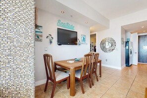 A dining area with a wooden table and chairs, a flat-screen TV mounted on the wall, and a decorative wall piece. - Emerald by the Sea #711 (Galveston)