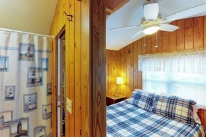 Una habitación con paredes de madera, una cama con colcha azul y blanca a cuadros, un ventilador de techo y una ventana con persianas.