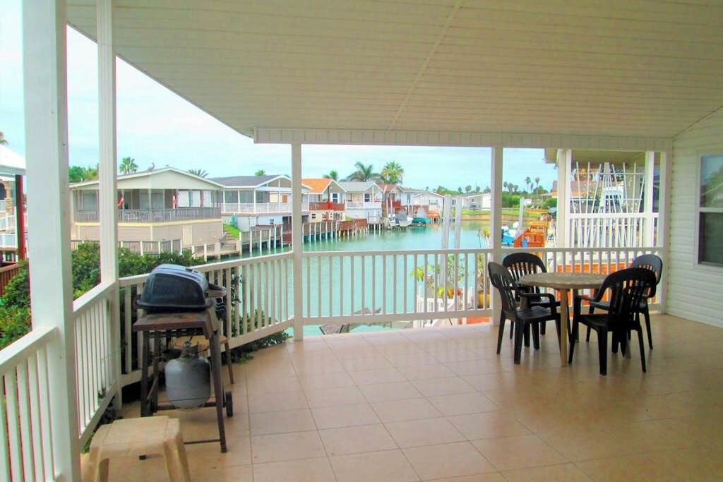 A covered outdoor area with a dining table and chairs, a grill, and a view of a water body with houses in the background.
