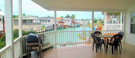 A covered outdoor area with a dining table and chairs, a grill, and a view of a water body with houses in the background.