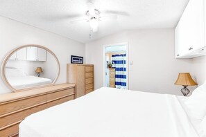 Quarto com cama, espelho redondo, guarda-roupa, ventoinha de teto e porta para um banheiro.