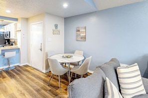 A modern kitchen with white cabinets, a dining table with chairs, and a gray sofa with striped pillows. - Beach Club 363 - Heated Pool & Hot Tub, Dogs OK (Corpus Christi)