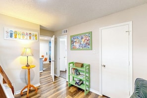 A hallway with a wooden floor, a green shelf with books, a lamp, and a door. - Beach Club 363 - Heated Pool & Hot Tub, Dogs OK (Corpus Christi)