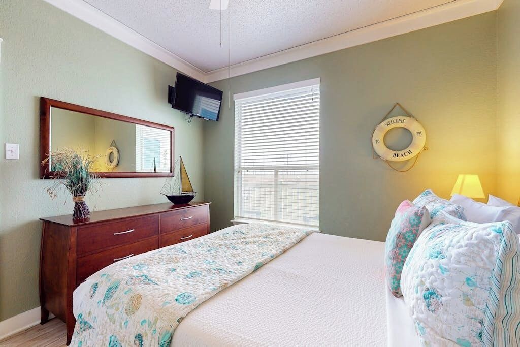 A bedroom with a bed, dresser, mirror, TV, window with blinds, and a decorative item on the wall.