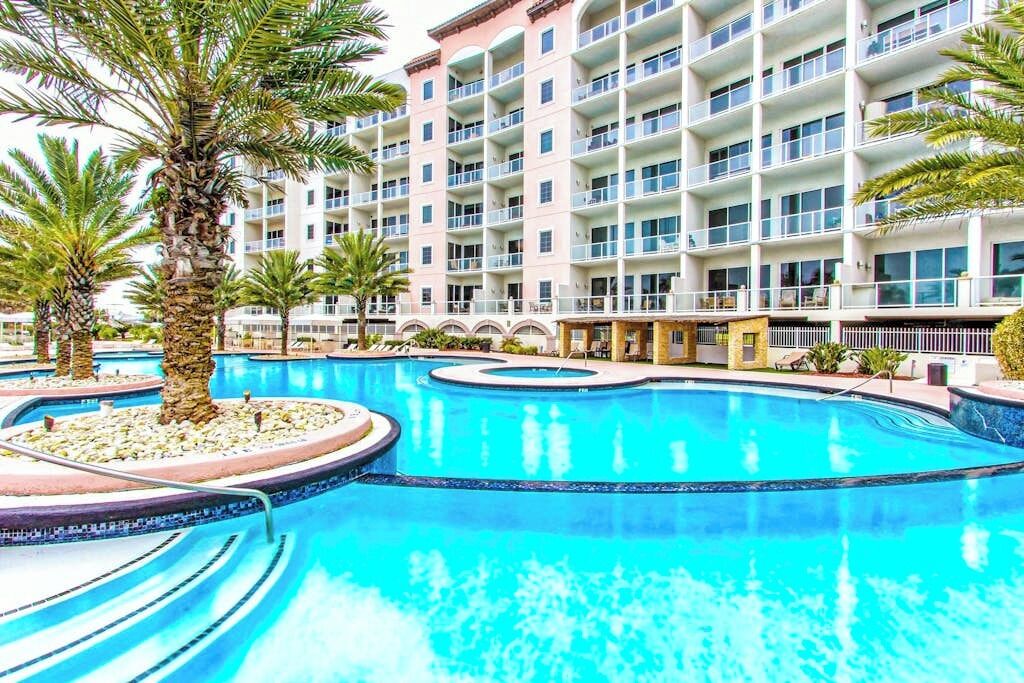 A hotel with a large swimming pool, palm trees, and a multi-story building with balconies.