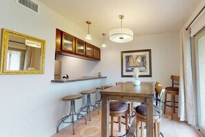 A dining area with a wooden table and bar stools, a kitchen with wooden cabinets, pendant lights, and a framed artwork on the wall. - Rockport Waterfront Getaway #608 (Rockport)