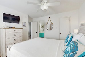 A bedroom with a bed, a ceiling fan, a television, a dresser, and a bathroom visible through an open door. - Beach Club 352 - Pool, Hot Tub, Gym & Balcony (Corpus Christi)
