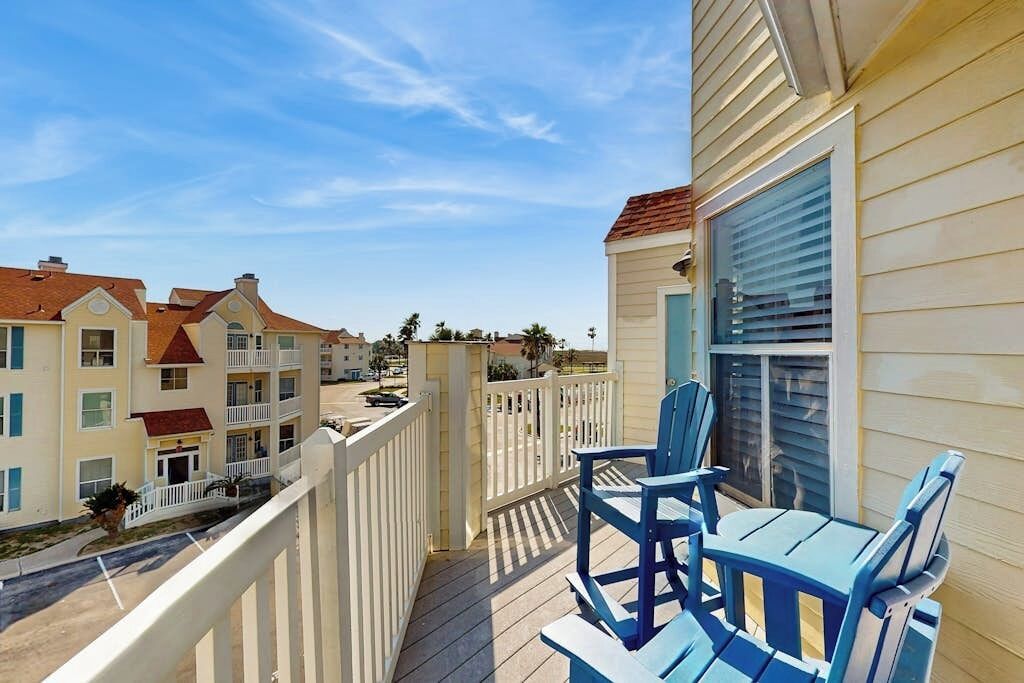 A balcony with blue chairs and a table, overlooking a street with multi-story buildings.